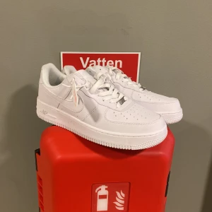 Nike Air Force 1 helvita sneakers - Nike Air Force 1 sneakers i helvitt med klassisk låg siluett. Skorna har snörning, perforerad tå och platt sula med rundad form. Tillverkade i skinn med syntetdetaljer och ikonisk swoosh på sidan. Perfekt för dig som gillar cleana och tidlösa sneakers.