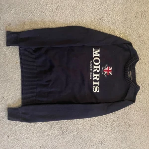 Mörkblå stickad tröja Morris XS - Snygg mörkblå stickad tröja från Morris med stor vit logotyp och broderad brittisk flagga på bröstet. Tröjan har ribbade muddar vid ärmslut och nederkant samt rund hals. Perfekt för dig som gillar klassisk stil med en modern twist.