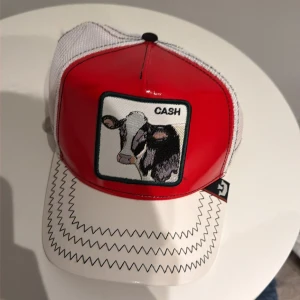 Röd och vit keps från The Farm - Snygg keps i rött och vitt med mesh på baksidan och justerbar snapback. Framtill finns ett broderat motiv med en ko och texten 'CASH'. Skärmen har zigzag-mönster och insidan är dekorerad med dollartecken. Perfekt för dig som gillar unika accessoarer.
