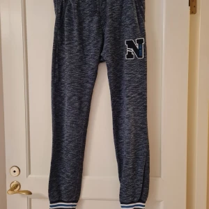 Mörkblå mjukisbyxor från Lindex strl 170 - Mörkblå mjukisbyxor från Lindex by Kids med melerad look och vit-blå rand på mudden vid benslut. Byxorna har resår i midjan och ett broderat N på ena benet. Storlek 170.