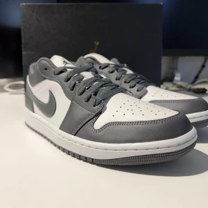 Nike Jordan 1 Low vit och grå- EUR 43- Helt Nya - Nike Jordan 1 Low sneakers i vit och mörkgrå färg med klassisk Swoosh på sidan.. Perfekt för dig som gillar stilrena och ikoniska sneakers. Skorna är helt nya, aldrig använda och kommer i original låda.
