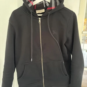 Svart Burberry hoodie - Svart hoodie från Burberry i storlek M med klassisk rutig insida på huvan i rött, grått och svart. Tröjan har dragkedja framtill, snörning vid huvan och två fickor. Broderad Burberry-logga på bröstet. Perfekt för dig som gillar stilrena streetwear-plagg.