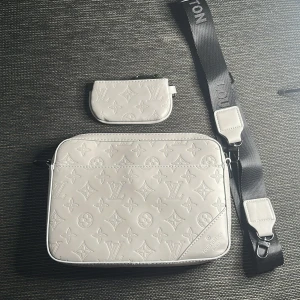 Vit axelväska - Snygg vit axelväska i skinn med klassiskt monogrammönster. Väskan har en bred svart axelrem och matchande liten plånbok. Perfekt för dig som vill ha en stilren och lyxig accessoar med ikonisk design.