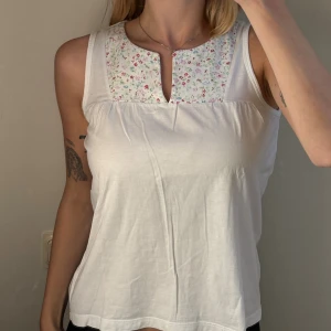 Vit blommig linnetopp från Jackpot - Supersöt vit linnetopp från Jackpot i mjuk bomull. Toppen har ett blommigt mönster upptill i rosa, lila och grönt, samt en liten v-ringad detalj vid halsen. Ärmlös och med avslappnad passform, perfekt för varma dagar. Säljer då den inte kommer till användning längre! 100% bomull