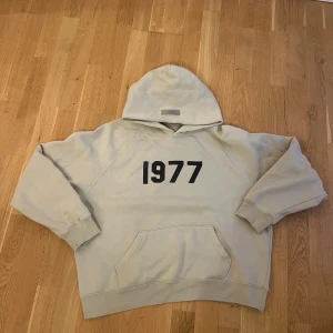 Beige mjukisbyxor Essentials Fear of God - Säljer ett par beige mjukisbyxor från Essentials Fear of God med trycket '1977' på vänster lår. Byxorna har elastisk midja med snörning och muddar vid bensluten. Materialet är mjukt och skönt, perfekt för chill dagar. Snygg streetwear-vibe och clean design.