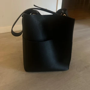 Svart axelväska i skinnimitation - Stilren svart axelväska med justerbart axelband och snygga gulddetaljer. Väskan har en fyrkantig form och är tillverkad i strukturmönstrad skinnimitation. Perfekt storlek för att få plats med allt du behöver till skolan eller stan.