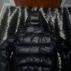 Svart dunjacka från Polo Ralph Lauren - Säljer en svart, glansig dunjacka från Polo Ralph Lauren med huva och dragkedja. Jackan har det klassiska röda Polo-broderiet på bröstet och är quiltad för extra värme. Perfekt för kalla dagar och har en riktigt snygg streetstil.