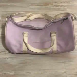Ljuslila weekendbag från Lacoste - Snygg ljuslila weekendbag från Lacoste med krämvita handtag och axelrem där Lacoste-loggan är broderad. Väskan har dragkedja upptill och är rymlig med innerfack. Perfekt för resor eller gymmet, stilren och praktisk design.