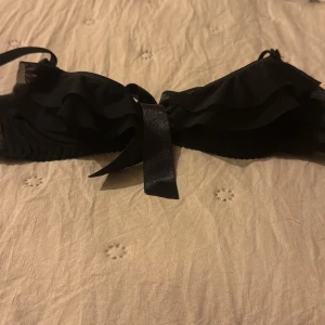 Svart bh med volang från Axami - Supersnygg svart bh från Axami med volangdetaljer och stor rosett framtill. Tunn mesh och glansigt band ger en lyxig känsla. Justerbara axelband och hakknäppning bak. 
