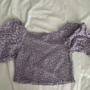 Lila blommig croppad blus med puffärm - Superfin lila blus med smockad kropp och croppad passform. Puffiga korta ärmar med små vita blommor och resår vid ärmslut.