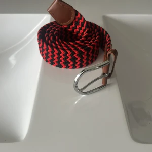 Randigt svart och rött flätat bälte - Snyggt flätat bälte i svart och rött med randigt mönster. Bältet har detaljer i brunt skinn vid ändarna och ett silvrigt metallspänne. Perfekt accessoar för att lyfta din outfit med lite färg och stil.