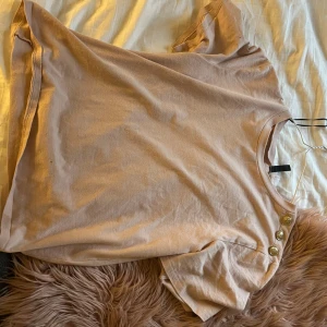 Beige kortärmad topp med knappar - Säljer en beige kortärmad topp från Divided i mjuk bomull. Toppen har rund halsringning och tre dekorativa guldknappar på ena axeln som snygg detalj. Perfekt för dig som gillar enkel men ändå unik stil. Pris går att diskutera 