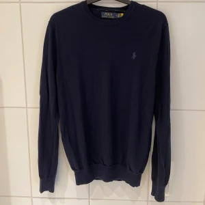 Mörkblå merinoulltröja Polo Ralph Lauren - Stilren marinblå tröja från Polo Ralph Lauren i slim fit-modell. Tillverkad i tvättbar merinoull, vilket gör den både bekväm och lättskött. Tröjan har den klassiska Polo-loggan broderad på bröstet i matchande färg.  Färg: Mörkblå / Navy Material: 100% tvättbar merinoull Passform: Slim fit Mått: Axelbredd 44 cm, längd 69 cm Skick: Mycket bra – sparsamt använd, utan fläckar eller hål  Perfekt till både vardag och mer uppklädda tillfällen.
