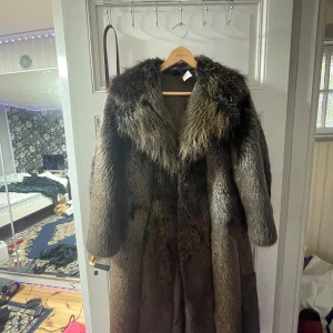 Lång brun pälsjacka med krage - Säljer en lång och maffig pälsjacka i mörkbruna och grå toner. Jackan har bred krage, rak passform och långa ärmar. Pälsen är tät och ger en riktigt lyxig känsla, perfekt för kalla dagar när du vill sticka ut.