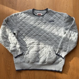 Grå quiltad sweatshirt från Levi's - Quiltad grå sweatshirt från Levi's med mörkblå broderad logga på bröstet. Tröjan har rund hals, ribbade muddar och en stor magficka. Materialet är mjuk bomull och polyester, perfekt för chilliga dagar. Snygga mörkblå detaljer vid hals och ärmslut.