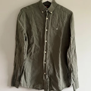 Skjorta  - Olivgrön skjorta från Morris i 100% linne. Klassisk modell med button-down krage, vita knappar och broderad logga på bröstet. Perfekt för dig som gillar en stilren och avslappnad look. Lite urtvättad 