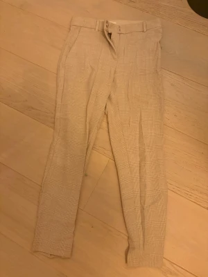 Beige rutiga kostymbyxor från H&M - Snygga beige kostymbyxor från H&M i storlek 40. Byxorna har ett rutigt mönster, smal passform och midwaist. De har bälteshällor, dragkedja och metallhakar i midjan. Perfekta för en clean och stilren look.