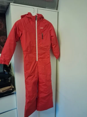 Röd täckoverall från Wedze, strl 130 - Röd täckoverall från Wedze med huva och vit dragkedja framtill. Overallen har en bröstficka med dragkedja och resår i midjan för bättre passform. Perfekt för kalla dagar och vinteraktiviteter. Märkt med Decathlon på insidan.