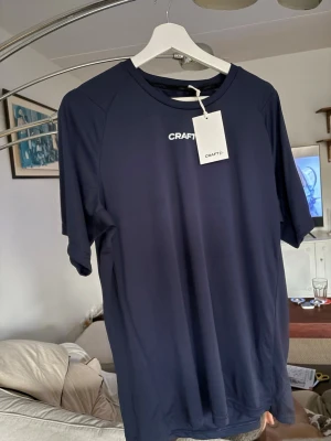 Mörkblå Craft t-shirt med tryck - Säljer en mörkblå t-shirt från Craft med vit logga framtill och stort tryck med motiv och texten 'Københavns Kommune' på ryggen. T-shirten har rund hals och är tillverkad i ett lätt syntetmaterial som passar perfekt för träning.