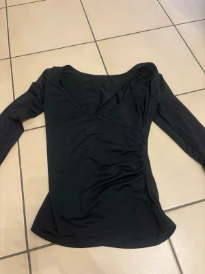 Svart omlottopp med lång ärm - Säljer en svart omlottopp med lång ärm och v-ringad hals. Toppen har snygga rynkade detaljer på ena sidan och är gjord i ett mjukt, stretchigt syntetmaterial. Perfekt för dig som gillar stilrena och enkla plagg med en twist.
