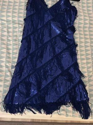 Blå paljettklänning med fransar - Säljer en cool blå kortklänning med glittriga paljetter och långa fransar i diagonala rader. Klänningen har v-ringad hals och är ärmlös, perfekt för dig som gillar att sticka ut. Materialet är polyester med paljettdetaljer och fransar som ger extra rörelse.