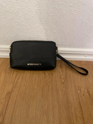 Svart handväska från Steve Madden - Snygg svart handväska från Steve Madden med guldfärgade detaljer och två dragkedjor. Väskan är tillverkad i strukturerat skinnimitation och har en praktisk handledsrem. Perfekt storlek för det viktigaste och stilren design med metallogo framtill.