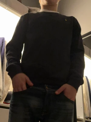Blå sweatshirt från Lyle & Scott - Blå sweatshirt från Lyle & Scott med klassisk rund hals och gul logotyp på bröstet. Tröjan har långärmade muddar och är gjord i mjuk bomull, perfekt för chill dagar. Enkel och stilren design som funkar till jeans eller joggers.
