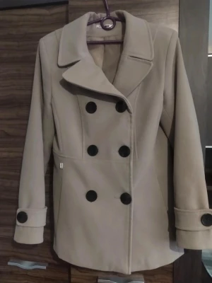 Beige skepparkavaj från Simge storlek 40 - Snygg beige skepparkavaj från Simge med stora svarta knappar fram och bak. Kappan har klassisk krage, långärmad design och är tillverkad i mjukt ullmaterial med slät insida. Perfekt för dig som vill ha en stilren och tidlös look. Kappan är i otroligt fint skick och knappt använd. Priset går att diskutera.