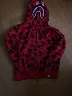 Röd Bape Hoodie  - Säljer denna feta röda bape haj-hoodie. Köpt från en grabb på Plick så är inte säker om den är äkta men den är i väldigt bra kvalité och är använd ett fåtal gånger. Säljer på grund av byte av stil! Hör av er vid frågor