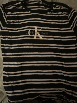 Svartvit randig t-shirt Calvin Klein - Säljer en svartvit randig t-shirt från Calvin Klein Jeans med CK-logga tryckt på bröstet. T-shirten har korta ärmar och klassisk rund hals. Materialet känns mjukt och skönt, perfekt för en chill och stilren look.