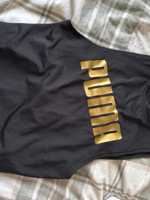 Svart linnetopp från Puma med guldtryck - Titta in på min sida för liknande. Säljer en svart linnetopp från Puma i storlek XS. Toppen har ett stort guldigt Puma-tryck framtill och snygg rynkdetalj på ryggen. Materialet är mjukt och stretchigt, perfekt för träning eller chill. Passformen är slim och modellen är ärmlös.