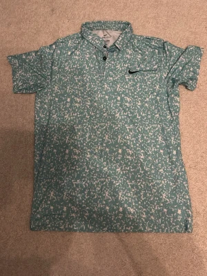 Golf Pike Nike Grön-vit M/L - Nike Dri-Fit skjorta i grön färg med vitt prickigt mönster. Piken har korta ärmar, klassisk krage och knappar framtill. Materialet är polyester som andas och känns lätt mot huden. Perfekt för dig som gillar sportig och fräsch stil och vill vara snyggast på golfbanan! Står storlek M men passar bättre för L
