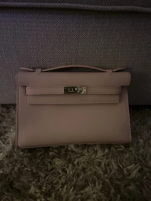 Pochette swift leather  - Säljer en stilren ljusrosa handväska i skinn med klassisk rektangulär form och elegant handtag. Väskan har snygga silverfärgade metalldetaljer och ett vridlås framtill. Perfekt för dig som gillar minimalistisk design och vill ha en chic accessoar.