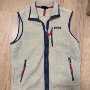 Beige fleeceväst från Patagonia - Säljer en beige fleeceväst från Patagonia med blå detaljer och röda dragkedjor. Västen har hög krage, tre fickor med dragkedja och klassisk Patagonia-logga på bröstet. Perfekt för lager-på-lager och outdoor-stil. Storlek M passar bra runt 190-185 på längden. Hör av er vid funderingar😄