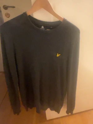 Mörkgrå långärmad tröja Lyle & Scott - Snygg mörkgrå långärmad tröja från Lyle & Scott med klassisk gul broderad fågel på bröstet. Tröjan har rund hals och är tillverkad i mjuk bomull, perfekt för chill dagar. Passar dig som gillar stilrena och enkla plagg med en touch av ikonisk detalj.