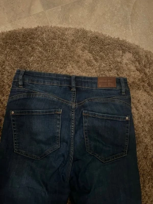 Mörkblå jeans från Ellos, storlek S - Snygga mörkblå jeans från Ellos med klassisk femficksdesign och raka ben. Jeansen har kontrastsömmar, bälteshällor och en brun läderpatch bak i midjan. Materialet är slitstarkt denim i bomull, perfekt för en chill och stilren look.