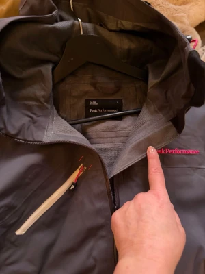 Grå Gore-Tex Peak Performance L - Peak Performance vindjacka i mörkgrått med rosa detaljer och logga. Jackan har huva, flera dragkedjeförsedda fickor och RECCO-reflektor på ärmen. Materialet är Gore-Tex och jackan är perfekt för utomhusaktiviteter. Snygg och funktionell design med sportig vibe.