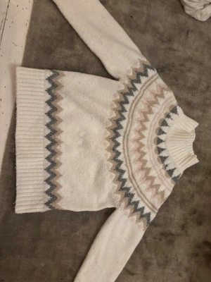 Vit stickad tröja med zigzagmönster, två små igen sydda hål på framsidan - Mysig vit stickad tröja med höghals och zigzagmönster i beige, grått och ljusrosa runt bröstet och ärmsluten. Tröjan har långa ärmar och bred ribbad mudd vid hals och nederkant. Perfekt för kalla dagar och ger en riktigt soft vintervibe.