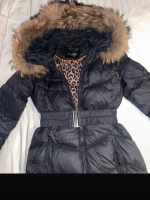Svart lång pufferjacka - Hollies  - Säljer en svart lång pufferjacka från Hollies med stor huva och brun päls. Jackan har bälte i midjan, dragkedja och knappar framtill samt praktiska fickor. Inga skador på jackan och alla knappar och dragkedjor fungerar. Fri från djur lukt och rökfri. 