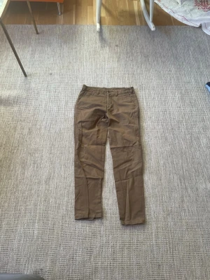 Bruna chinos från ARKET - Säljer ett par bruna chinos från ARKET med klassisk rak passform och midwaist. Byxorna har bälteshällor, sidofickor och bakfickor med knapp. Materialet är mjuk bomull som känns skönt mot huden. Perfekt för dig som gillar stilrena och enkla byxor.