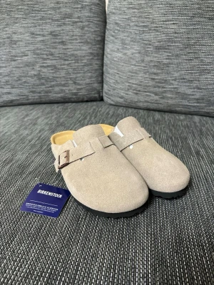 Beige Birkenstock Boston i mocka - Birkenstock Boston i beige mocka med justerbart spänne i sidan. Skorna har en klassisk korksula och svart yttersula med mönster. Perfekta att glida i och ur, med en stilren och enkel design som passar till många outfits.