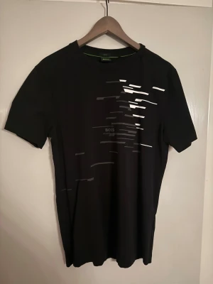 Svart t-shirt från BOSS med tryck - Snygg svart t-shirt från BOSS med coolt grafiskt tryck i vitt och grått på framsidan. T-shirten har rund hals och korta ärmar, perfekt för en stilren och modern look. Materialet känns mjukt och skönt mot huden.
