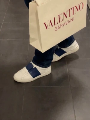 Valentino Garavani Open sneakers vit/blå - Säljer ett par Valentino Garavani Open sneakers i vitt skinn med bred marinblå rem över vristen. Klassisk rund tå, snörning och coola nitar på hälen. Kommer med originalkartong, dustbag och påse. Perfekta för dig som vill ha en lyxig och stilren sneaker. Aldrig använda!