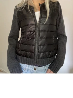Svart och grå pufferjacka med stickade ärmar - SÅ SNYGG CARDIGAN har i storlek 36,38 och 40❤️ på bilden är de storlek 36! Snygg pufferjacka i svart med grå stickade ärmar och ribbad kant. Jackan har dragkedja framtill och två fickor. Kroppsdelen är quiltad och ger en cool kontrast mot de mjuka stickade partierna. Perfekt för dig som gillar streetstyle och vill ha något unikt.