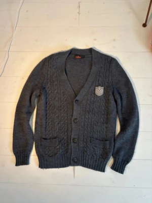 Grå Morris Merino Cardigan - Säljer en mörkgrå stickad kofta från Morris med snyggt kabelmönster och ett broderat emblem på bröstet. Koftan har v-ringning, knappar framtill och två fickor. Perfekt för lager-på-lager under höst och vinter.