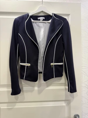 Svart kavaj med vita detaljer från H&M - Kavaj från H&M med vita kantband och guldfärgade dragkedjor vid fickorna. Jackan har en figurnära passform. Den är i storlek 36 men den passar mer 34 då den är liten i storleken. 