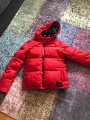 Röd pufferjacka från Calvin Klein Jeans - Säljer en röd pufferjacka från Calvin Klein Jeans med huva och svart innerfoder. Jackan har en rak passform, dragkedja framtill och ett stilrent märke på nederkanten. Perfekt för kalla dagar och riktigt snygg till streetwear. Det står att det är för 16 år gamla, men passar också för st 36 alltså S. Det finns en mindre fläck på baksida och mini hål som knabbt syns.