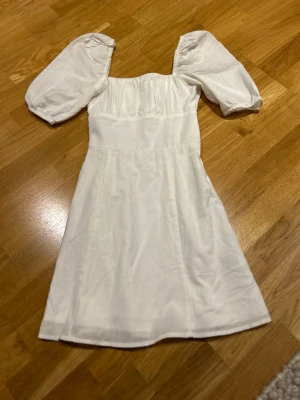 Vit kortklänning med puffärm XS H&M Divided - Säljer en vit kortklänning från H&M Divided i storlek XS. Klänningen har puffiga kortärmar och en smickrande fyrkantig halsringning. Tillverkad i ett lätt och mjukt bomullsmaterial som känns skönt mot huden. Perfekt för varma dagar, skolavslutning och snygg att styla med sneakers eller sandaler.
