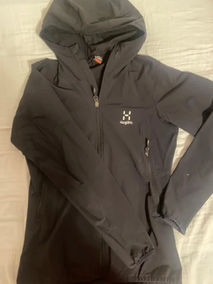 Svart vindjacka från Haglöfs, S - Svart vindjacka från Haglöfs i storlek S, med huva och hel dragkedja framtill. Jackan har två sidofickor med dragkedja och diskret logga på bröstet. Perfekt för blåsiga dagar och outdoor-aktiviteter. Snygg och funktionell design som passar dig som gillar friluftsliv. (se bild 3,5 för defekter)