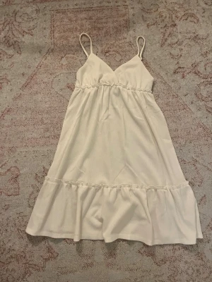 Vit omlottklänning från H&M - Supersöt vit klänning med tunna axelband och omlott-v-ringning från H&M. Klänningen har volangkant nedtill och är lätt och luftig, perfekt för varma dagar. Materialet känns mjukt och har en lätt struktur. Passformen är normal och modellen är kort.